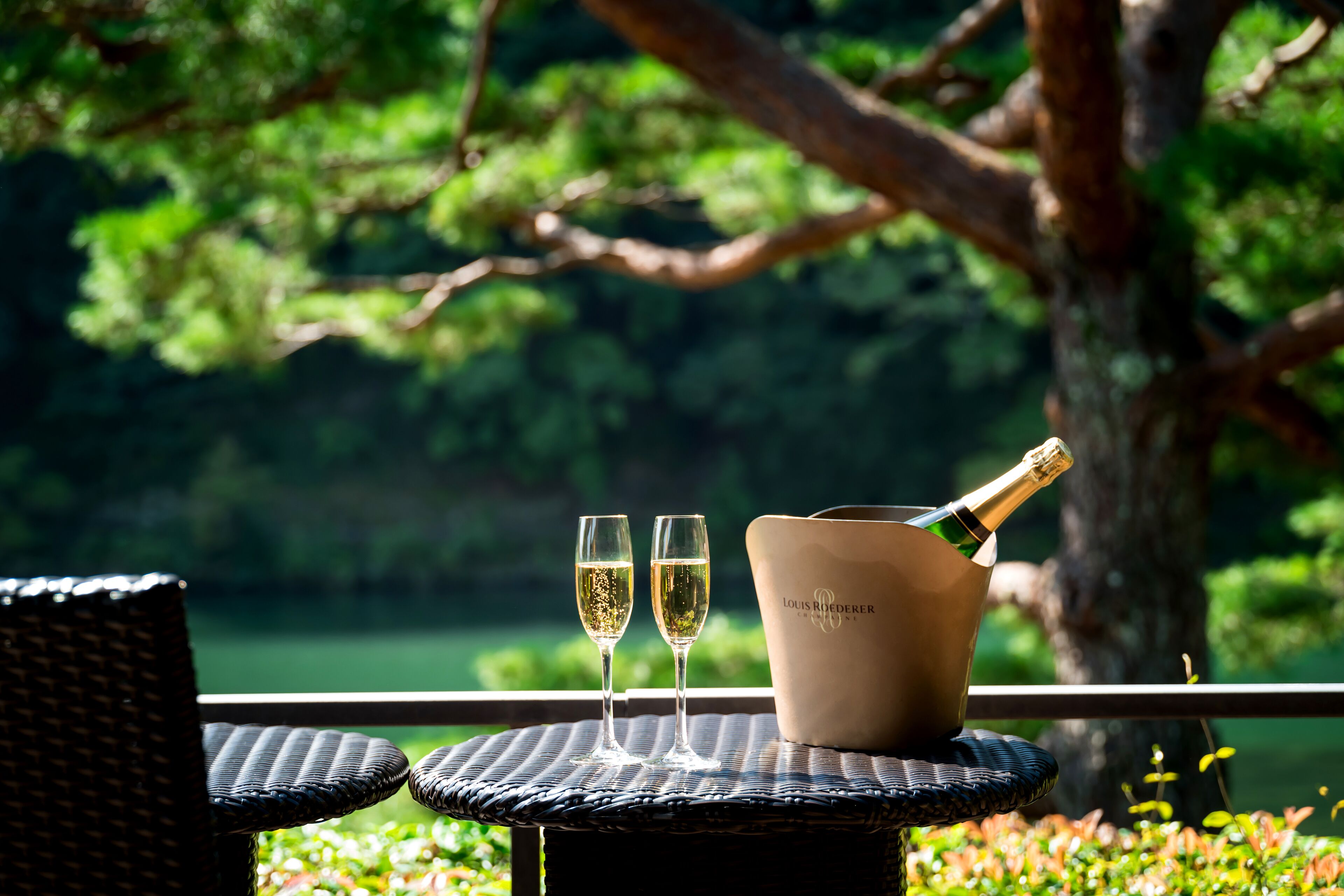 A wicker table with two glasses of champagne and a bottle on a balcony overlooking a green landscape.