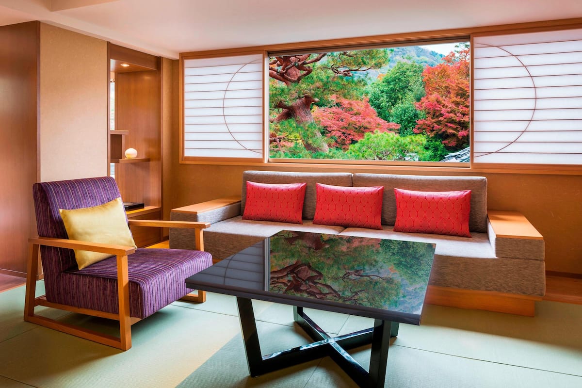 A traditional Japanese-style room with tatami flooring, a low table, a sofa with red cushions, and a view of a forest through shoji screens.