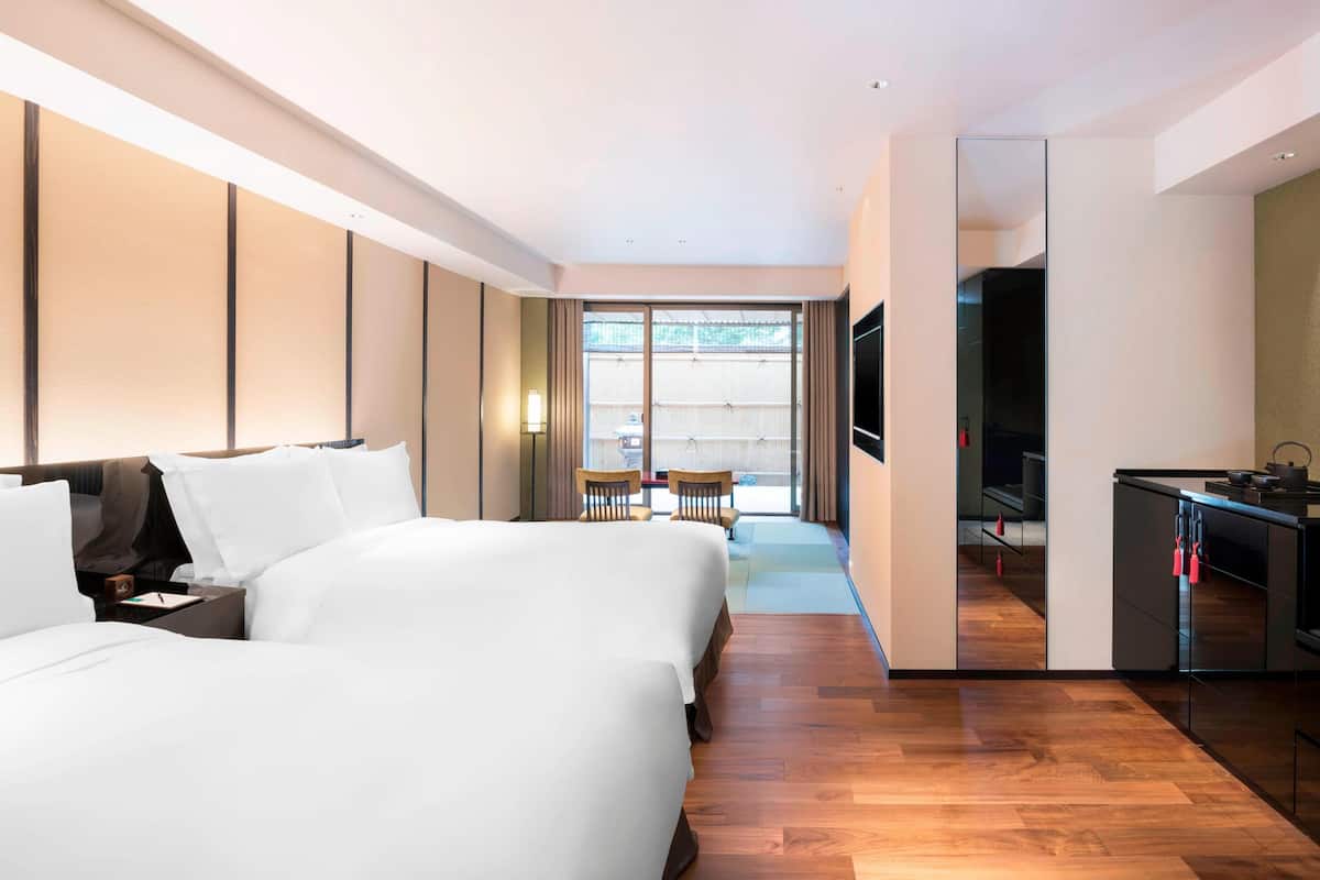 A hotel room with two beds, a TV, a kitchenette, and a balcony with chairs.