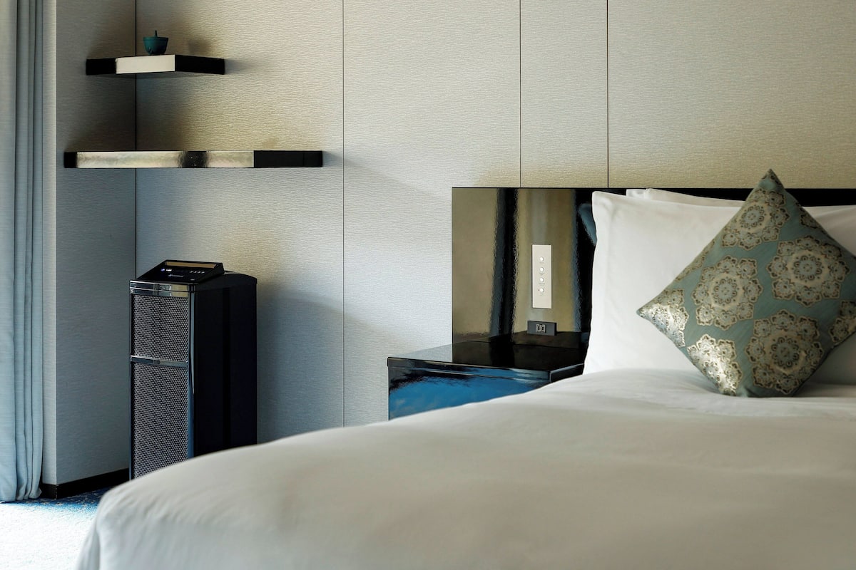 A hotel room with a bed, a nightstand, a mirror, a shelf with a bowl, and a black speaker.