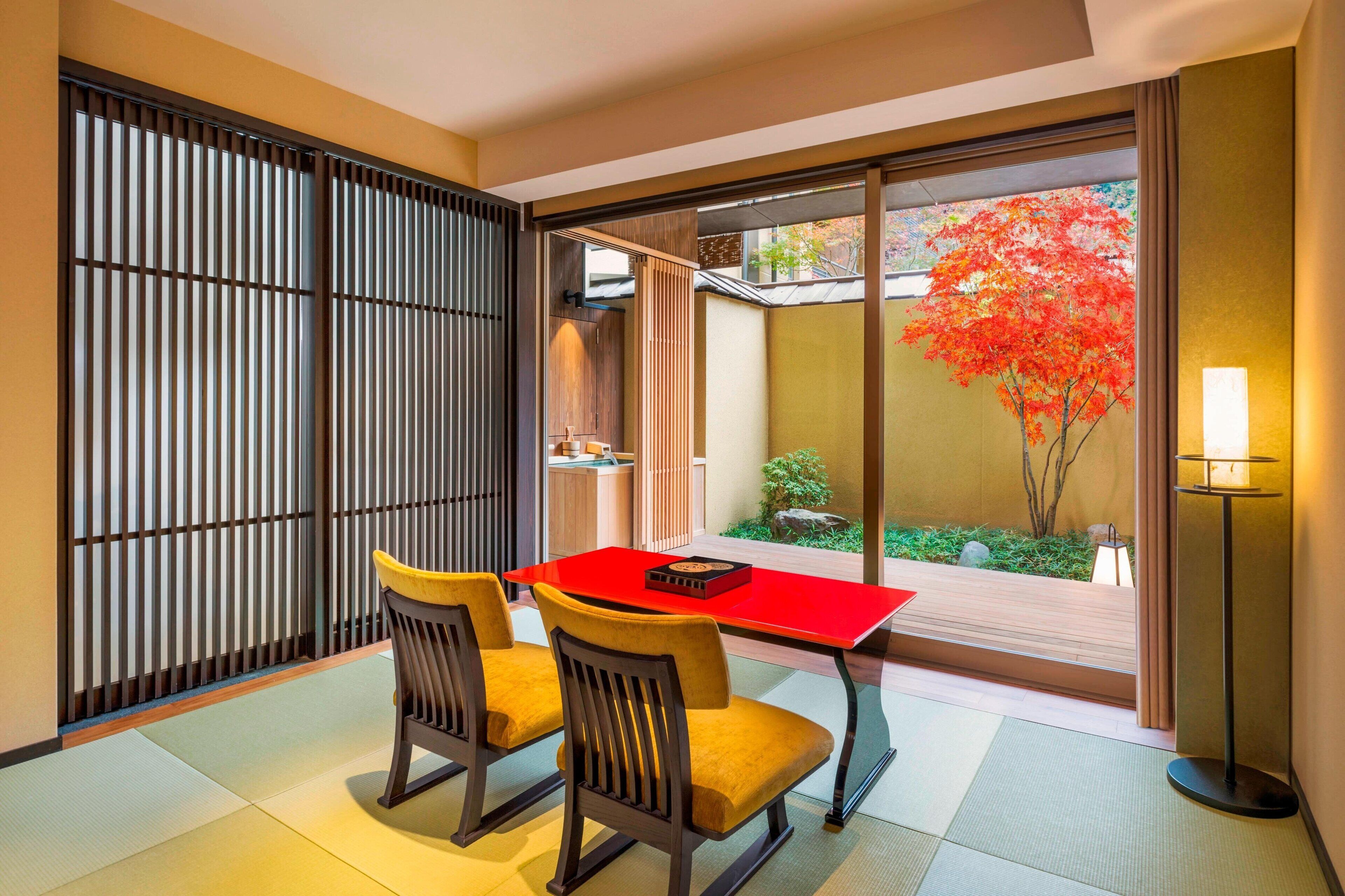 A traditional Japanese room with tatami flooring, a low table with two chairs, a sliding shoji screen, and a view of a garden with a red maple tree.