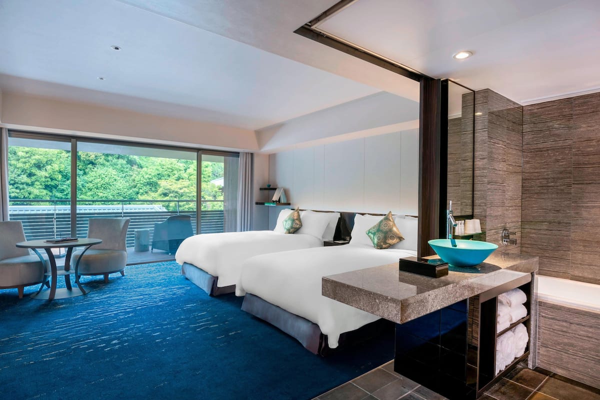 A modern hotel room with two beds, a large window, a balcony, and a bathroom with a blue sink.