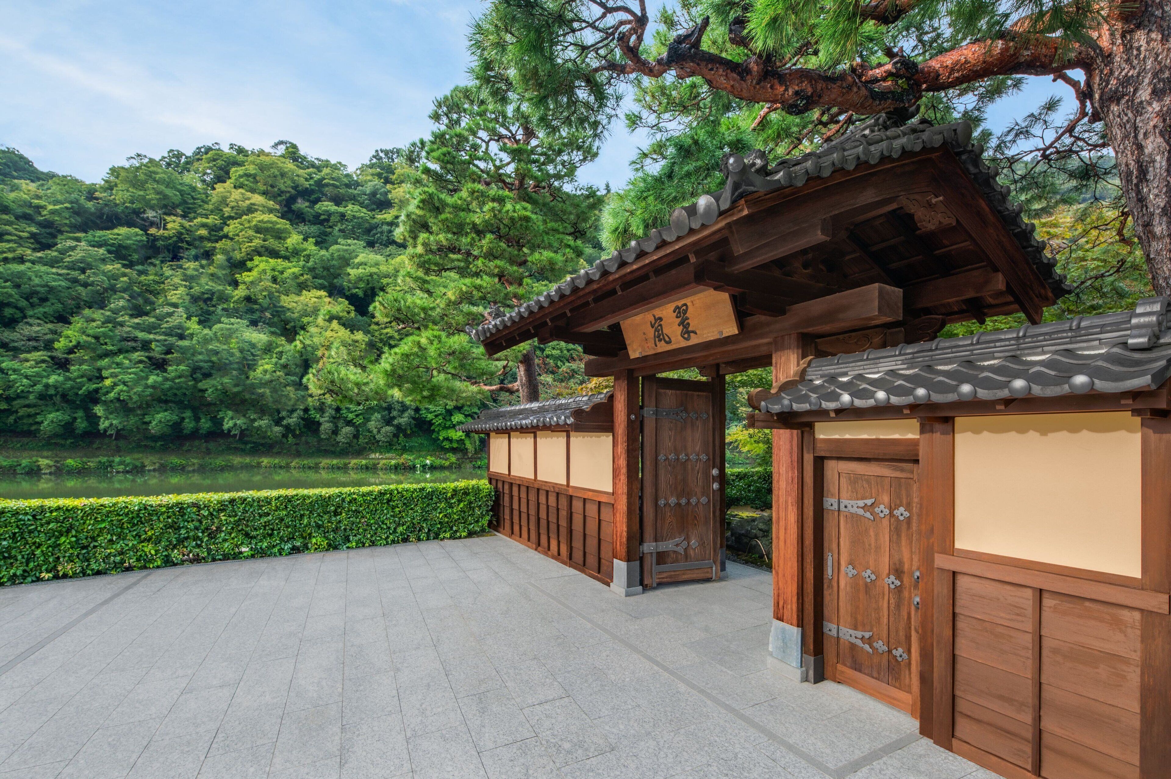A traditional Japanese gate with wooden architecture and a signboard, surrounded by lush greenery and a pond.