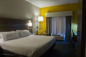 Quarto de hotel com cama grande, mesa de cabeceira, luminária, cadeira, ar condicionado e janela com cortinas.