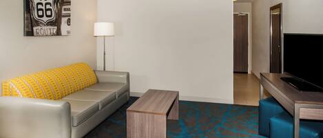 Une chambre d’hôtel moderne avec un canapé gris, une table basse en bois, un téléviseur et un tapis à motifs.