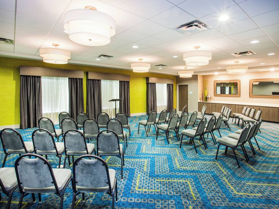 Un salón de conferencias con filas de sillas, un amplio ventanal con cortinas y suelo alfombrado.
