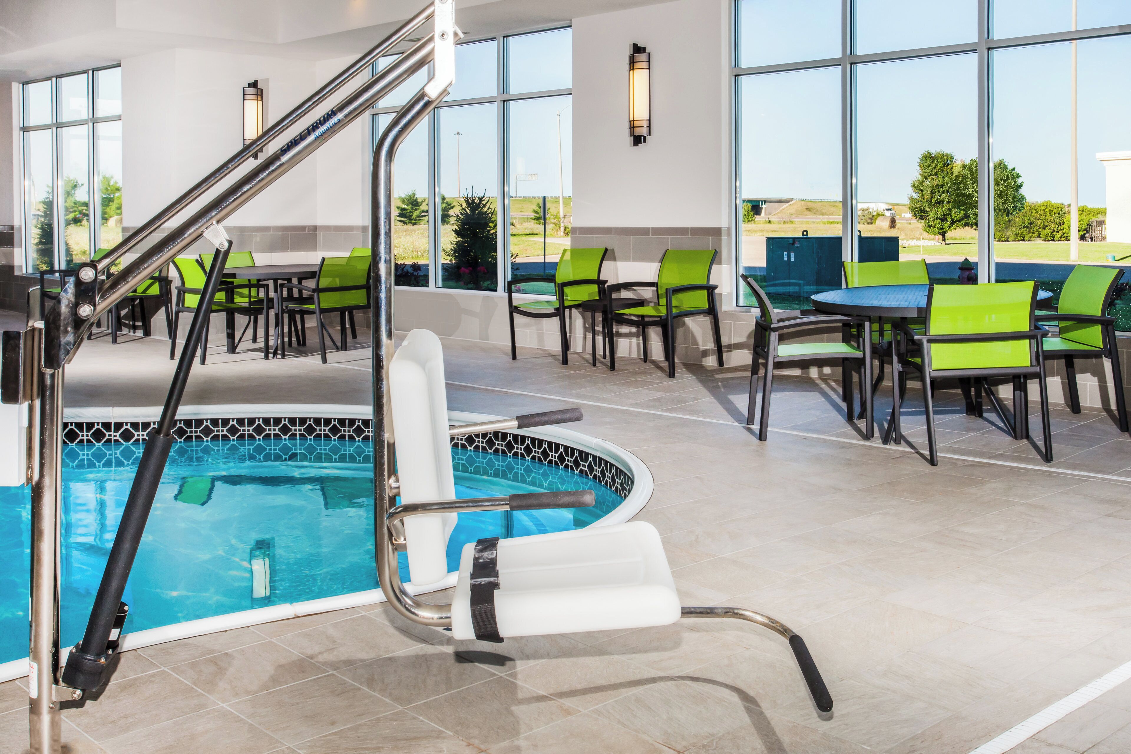 An indoor pool area with a pool ladder, green and black chairs, and a view of the outdoors through large windows.