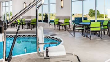 An indoor pool area with a pool ladder, green and black chairs, and a view of the outdoors through large windows.