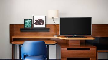 A modern hotel room with a wooden desk, a blue chair, a flat-screen TV, a lamp, and framed artwork on the wall.
