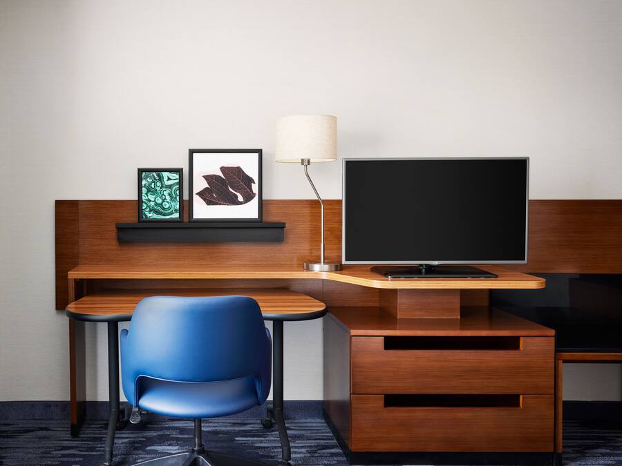 A modern hotel room with a wooden desk, a blue chair, a flat-screen TV, a lamp, and framed artwork on the wall.