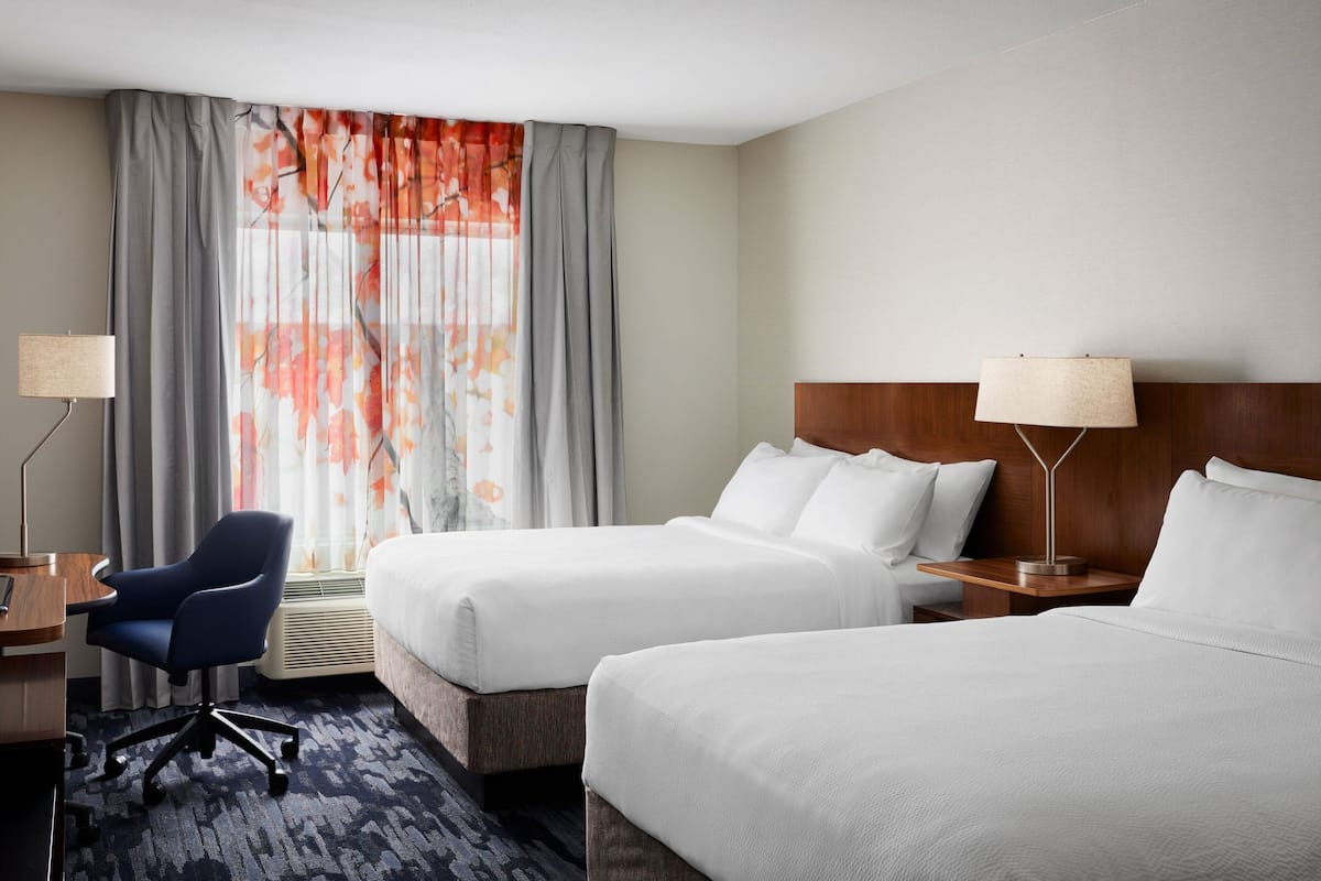 Une chambre d’hôtel avec deux lits, un bureau avec une chaise, une lampe et une fenêtre avec des rideaux.