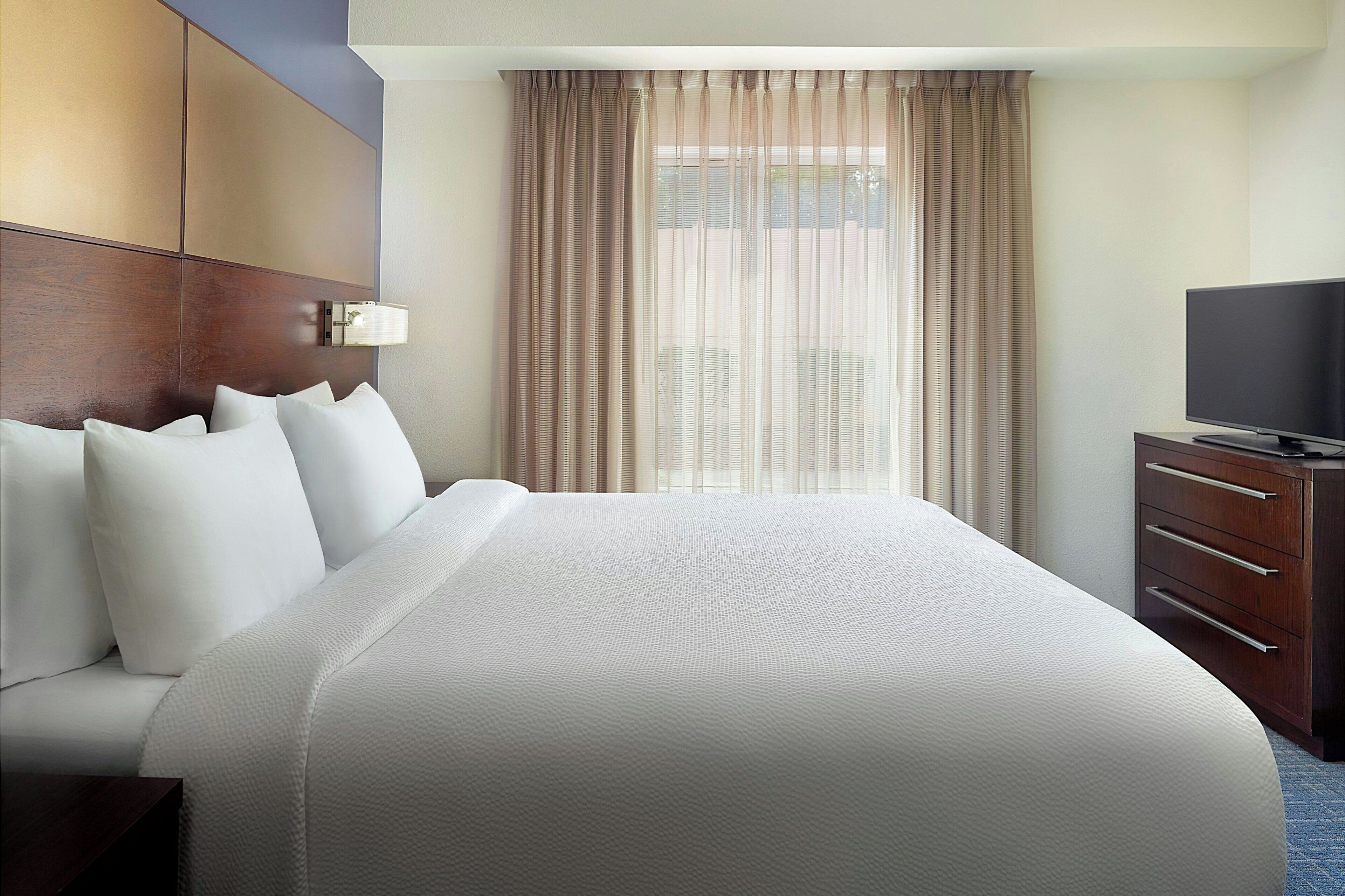 A hotel room with a large bed, a television on a wooden dresser, and a window with curtains.