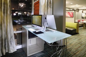 Habitación de hotel con un escritorio de vidrio, dos monitores, un ordenador de sobremesa y un sofá.
