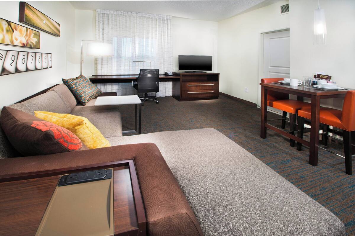 Habitación de hotel con sofá, escritorio con computadora, televisión y mesa de comedor con sillas.