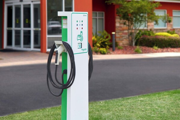 Una estación de carga pública para vehículos eléctricos, con una combinación de colores verde y blanco, ubicada frente a un edificio de fachada roja y entrada de vidrio.