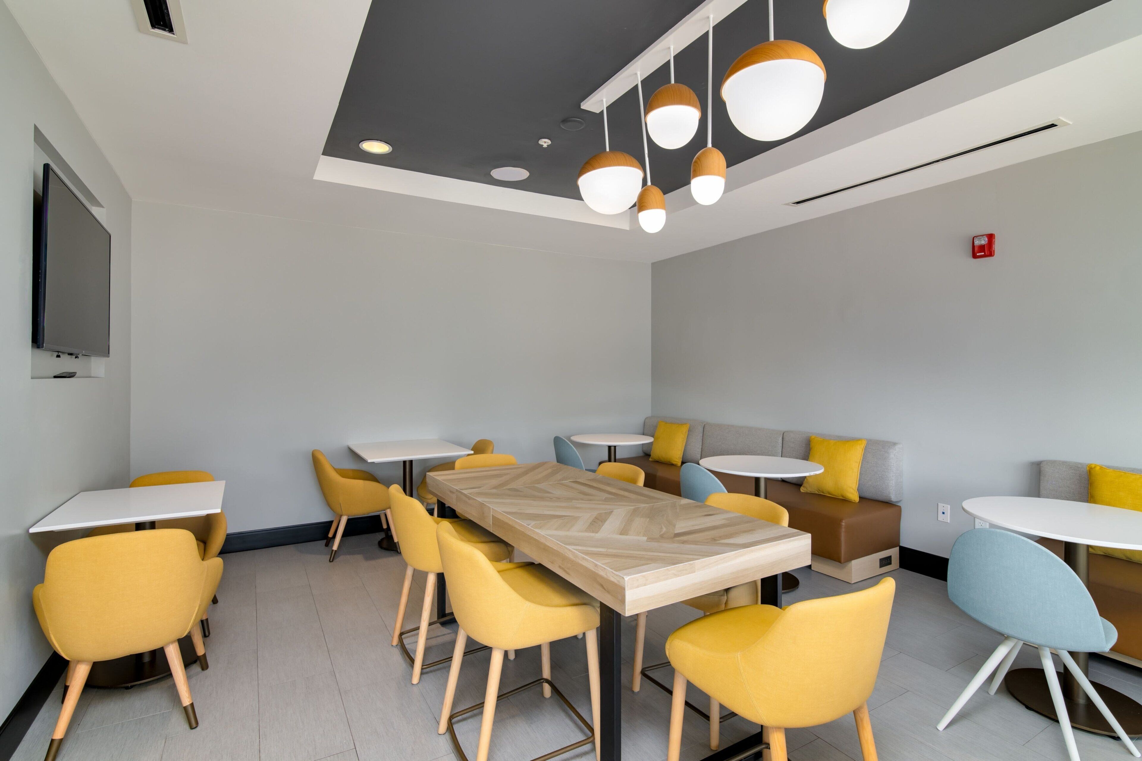 A modern meeting room with a long table and yellow chairs, a TV mounted on the wall, and a ceiling with pendant lights.