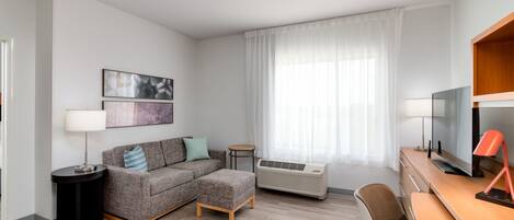 A modern hotel room with a grey sofa, a small round table, a desk with a computer, and a large window with sheer curtains.