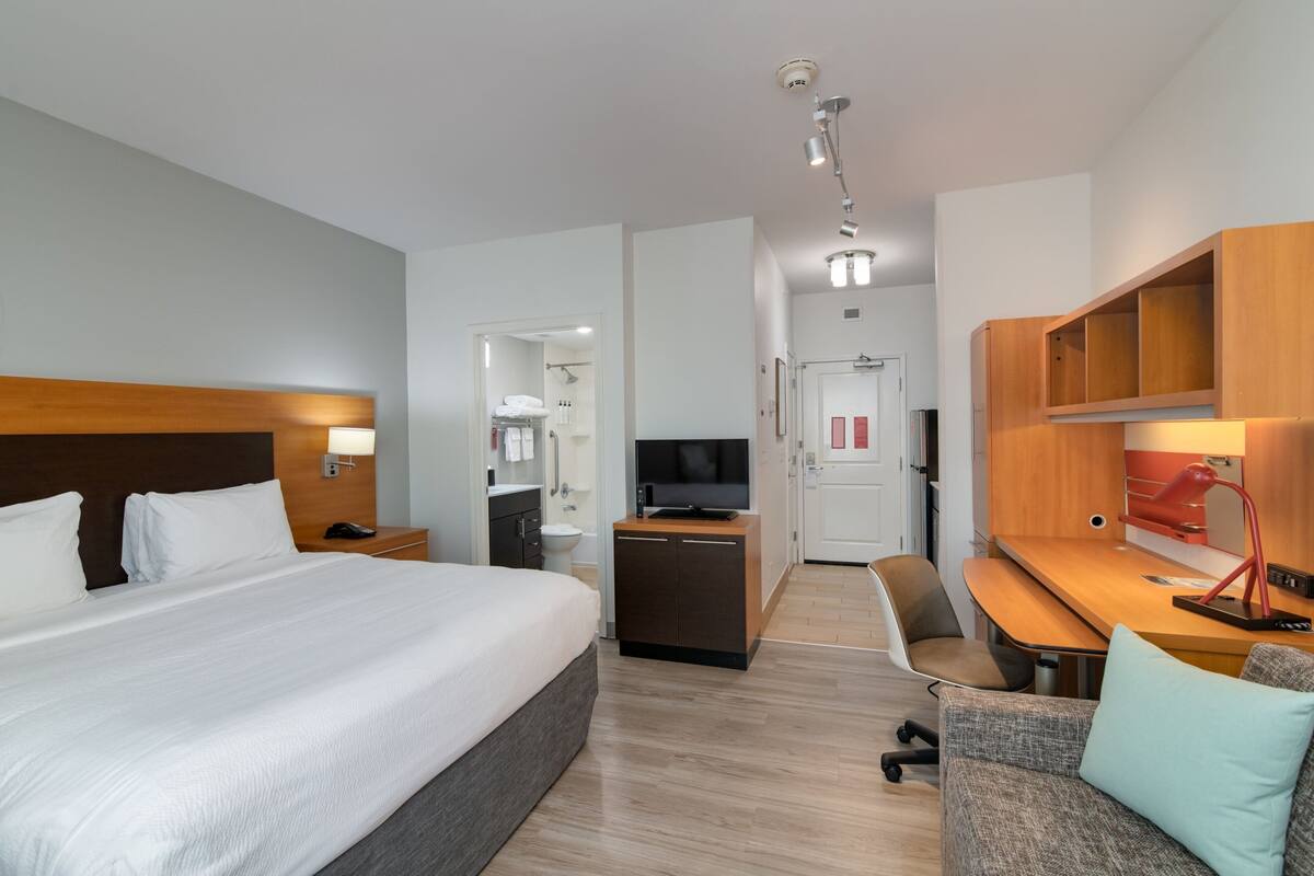 A hotel room with a large bed, a desk, a chair, a TV, and a bathroom visible through an open door.