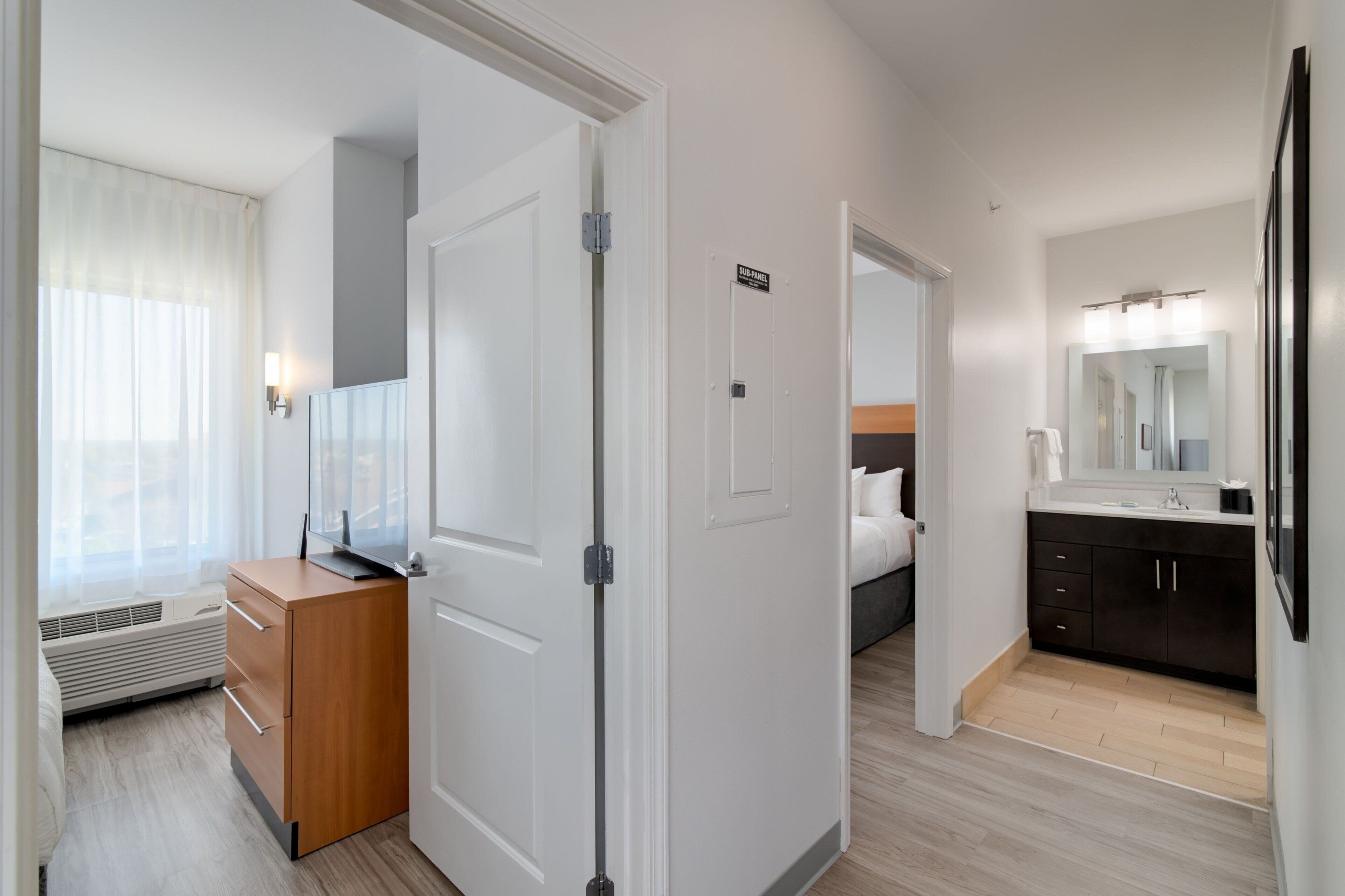 A hotel room with a wooden dresser, a bed, a bathroom with a dark vanity and mirror, and a hallway with a door.