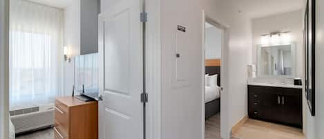 A hotel room with a wooden dresser, a bed, a bathroom with a dark vanity and mirror, and a hallway with a door.