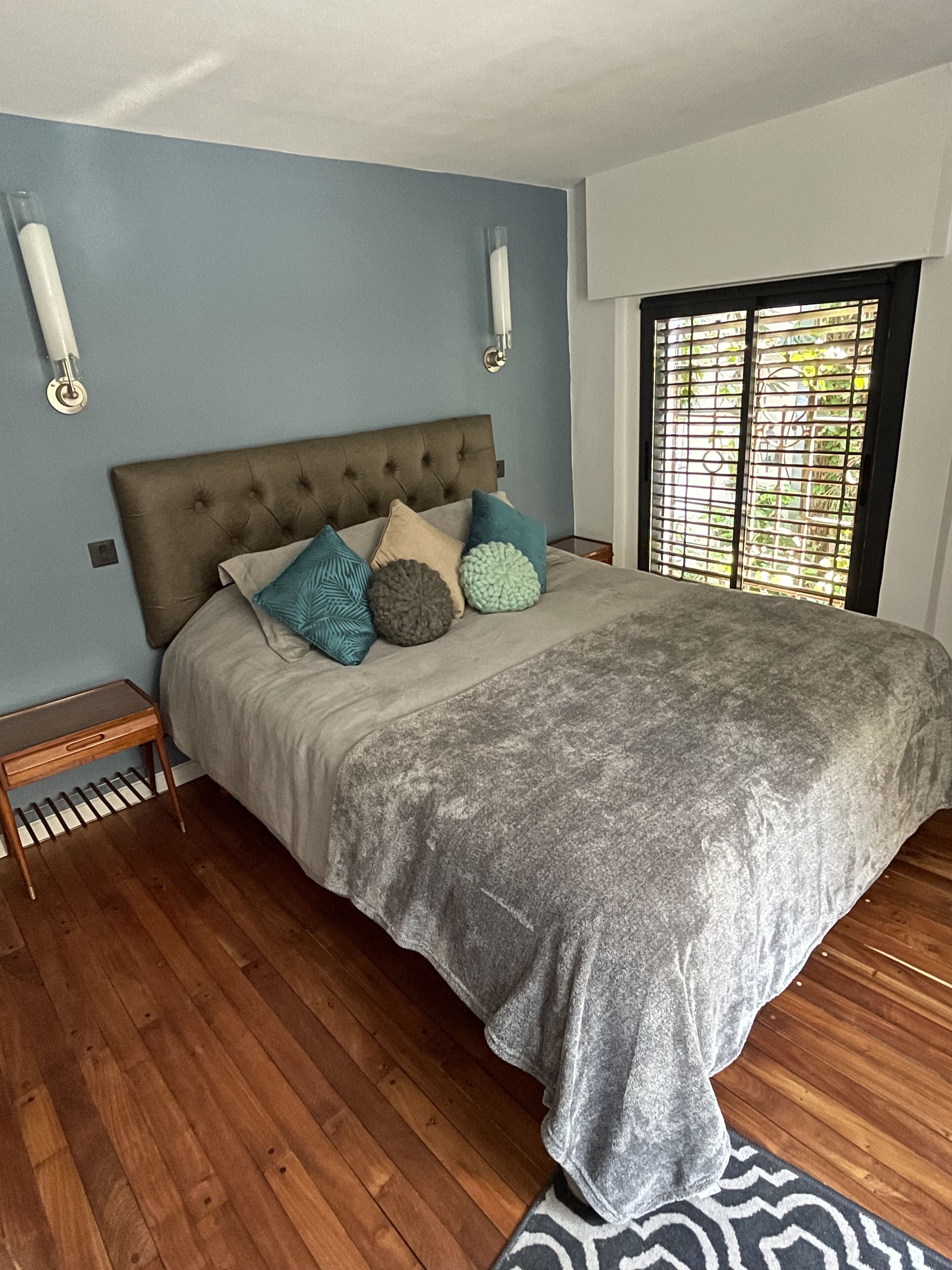 A bedroom with a tufted headboard, a grey bedspread, and multiple pillows. There is a wooden nightstand and a patterned rug.
