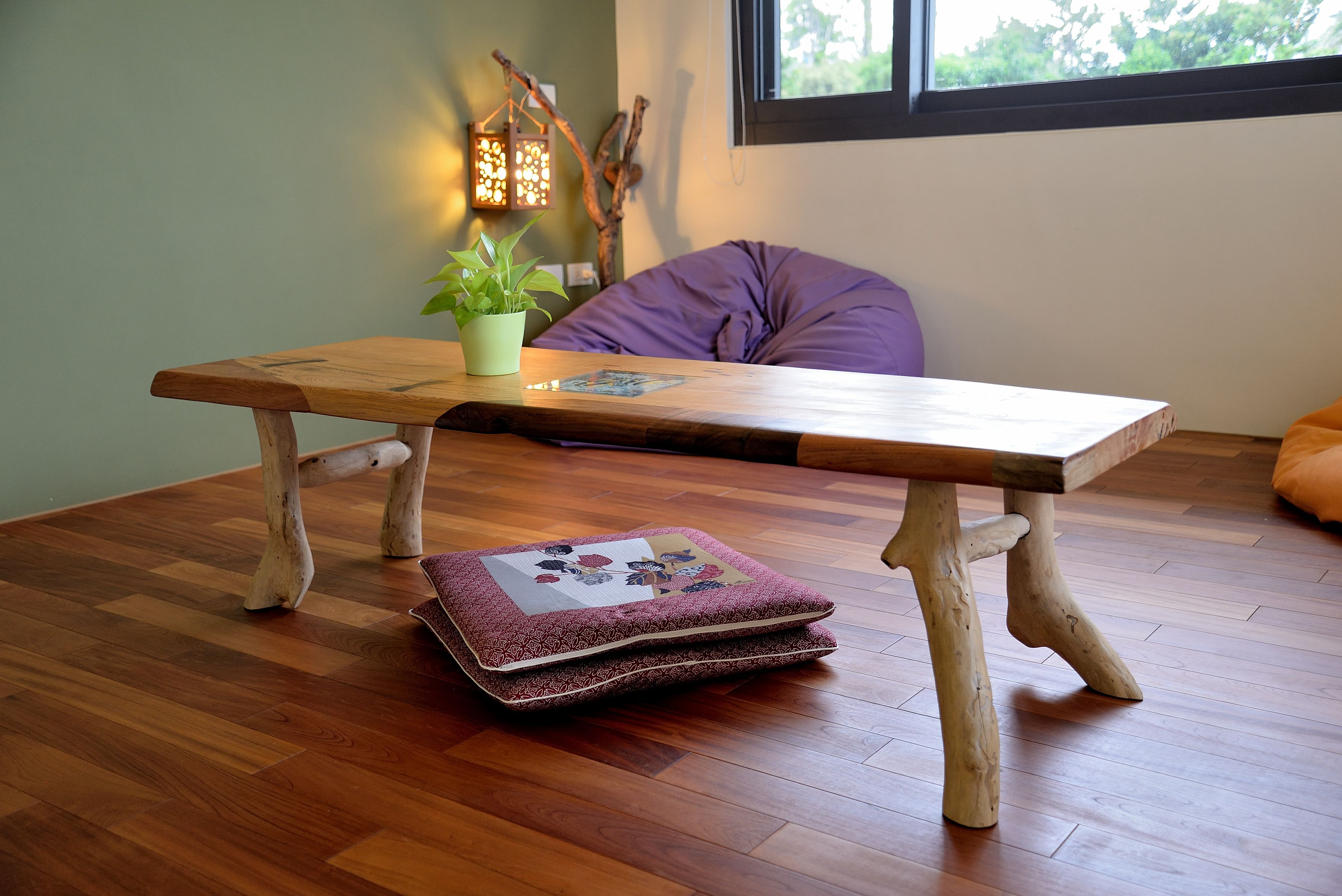 Una mesa de madera con un cojín y una planta encima, un cojín morado, y una lámpara colgante.