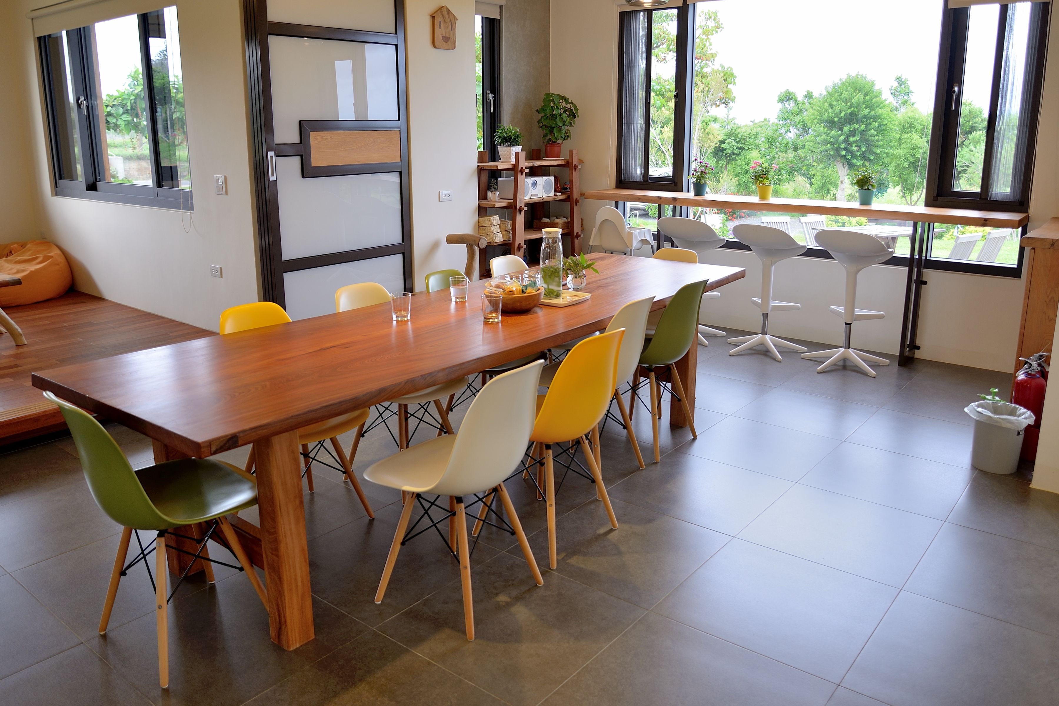 A dining area with a wooden table, multiple chairs, a bookshelf, and a view of greenery outside.