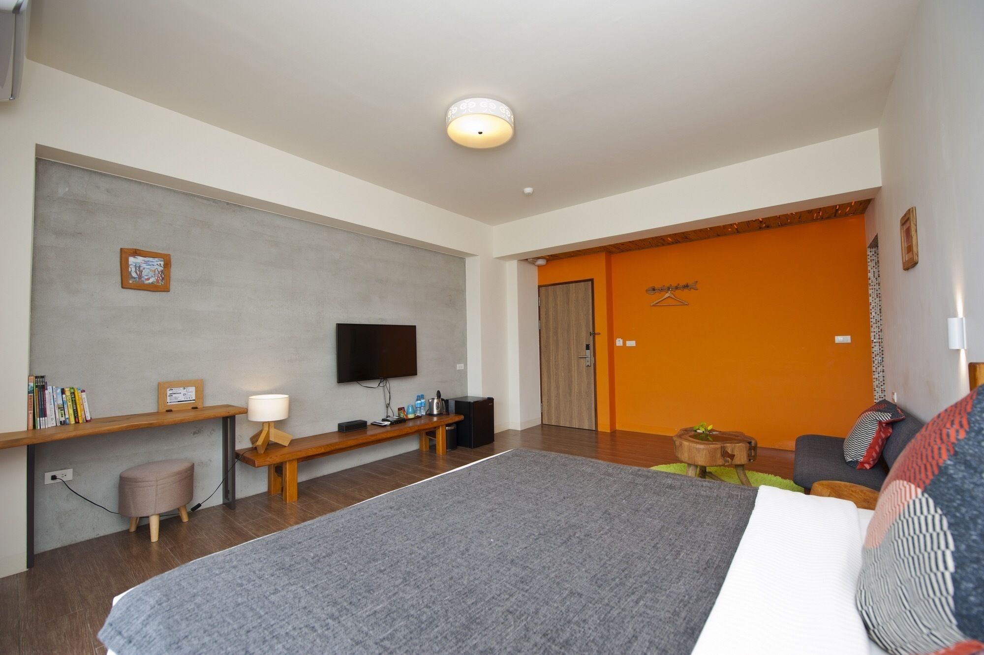 A modern hotel room with a large bed, a flat-screen TV mounted on the wall, a wooden bench, a small stool, and an orange door.