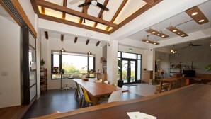 Un vaste espace dédié à la salle à manger, avec une longue table en bois, de nombreuses chaises et des luminaires suspendus. De grandes fenêtres laissent entrer la lumière naturelle et offrent une vue sur l’extérieur.