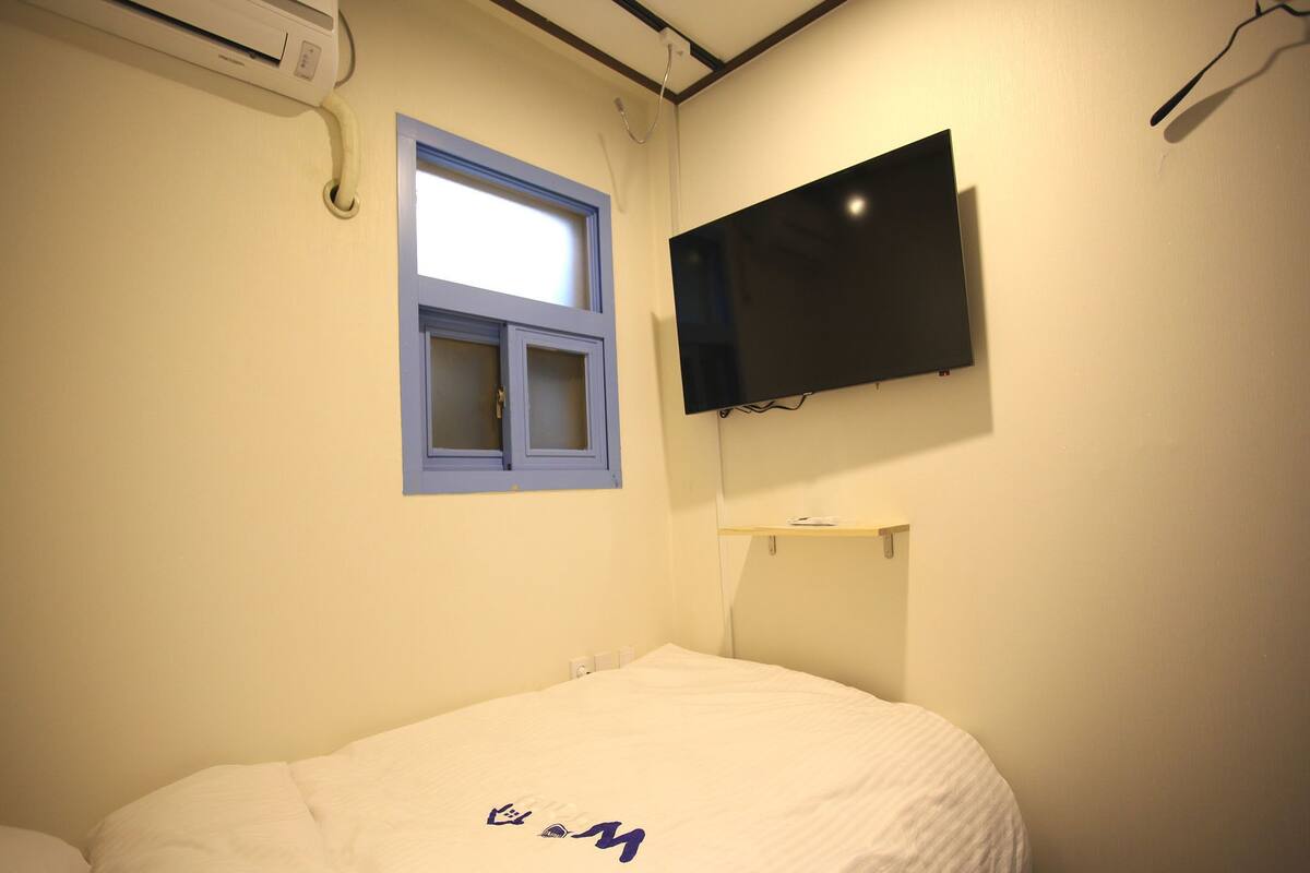 Una habitación con cama, ventana, televisor de pantalla plana y estante fijado a la pared.