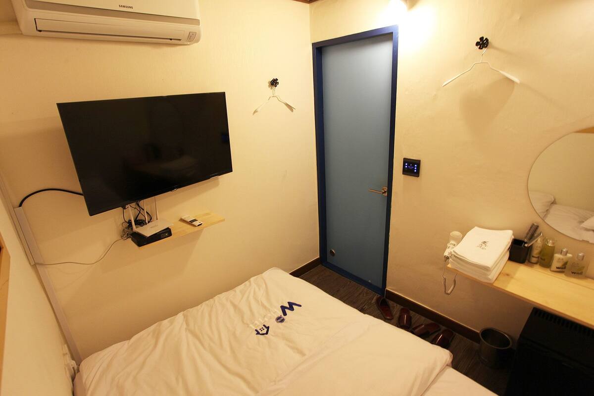 Habitación de hotel con cama, televisión montada en la pared, puerta, espejo y estante con artículos de aseo.