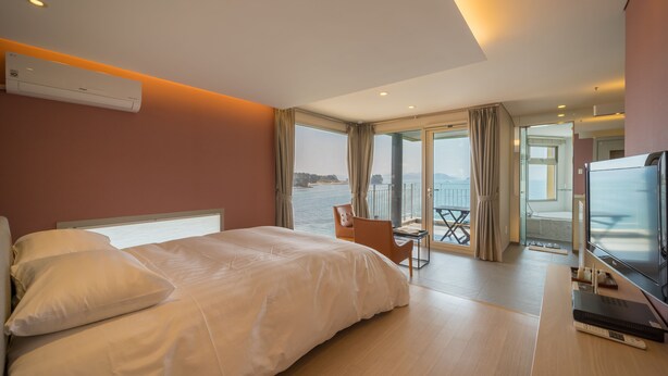 A modern hotel room with a large bed, a desk with a chair, a television, and a balcony with a view of the sea. - Tacet Pension (Tongyeong)