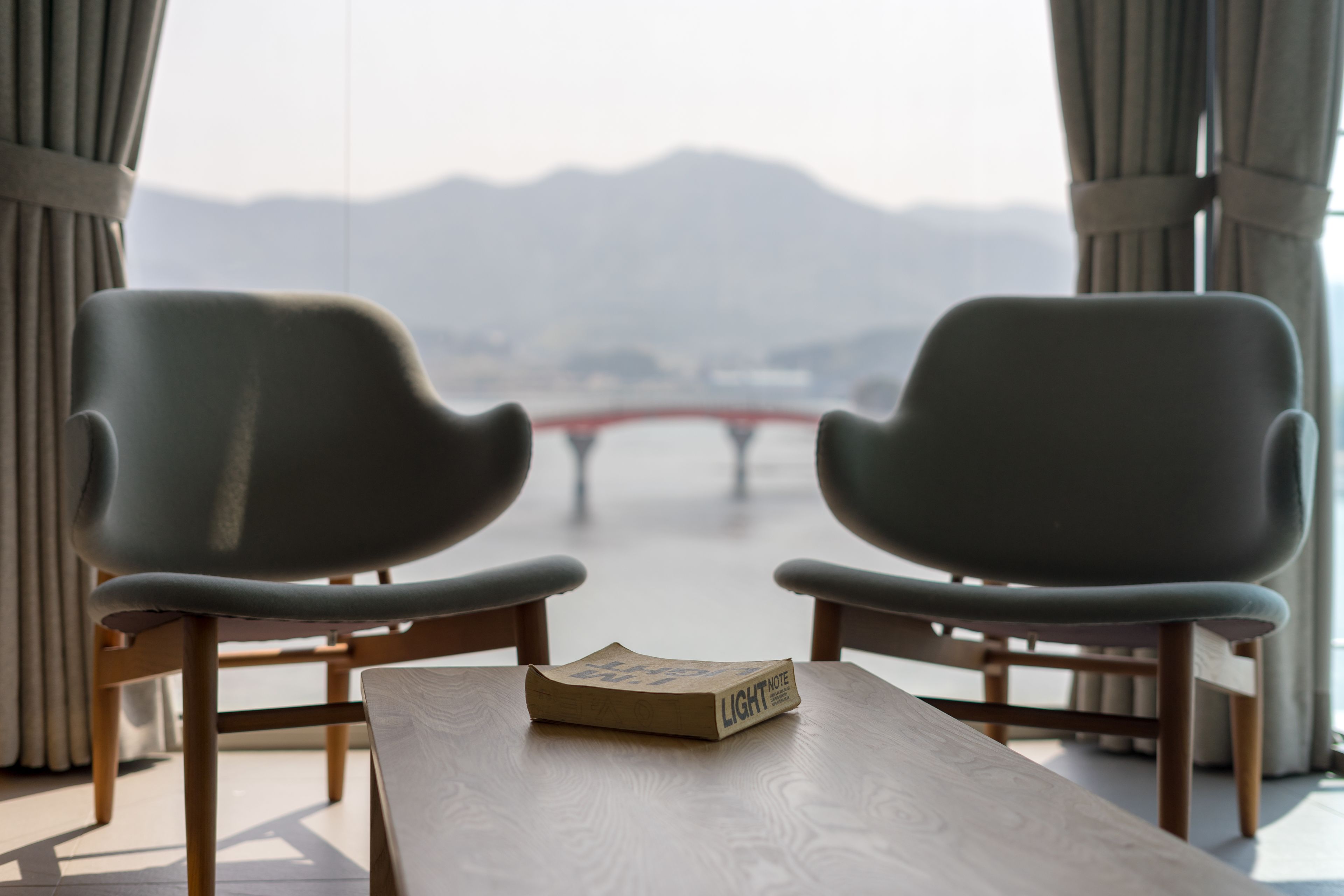 Duas cadeiras de frente para uma mesa com um livro intitulado "LIGHT NOTE". No quarto, vista para montanhas através de uma janela.