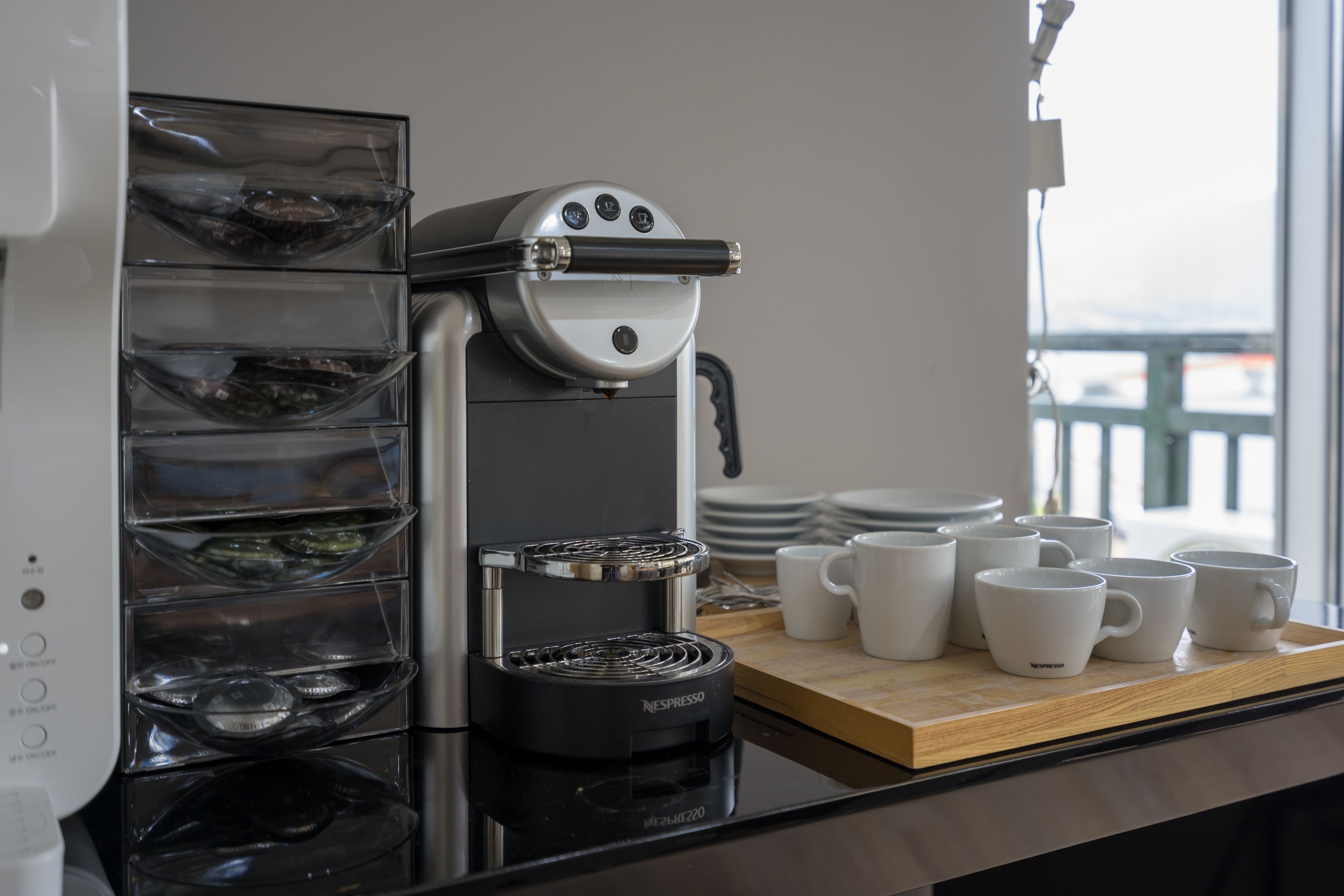 Una máquina de café Nespresso con jarra de vidrio y bandeja de goteo, una bandeja de madera con tazas blancas y una pila de platos.