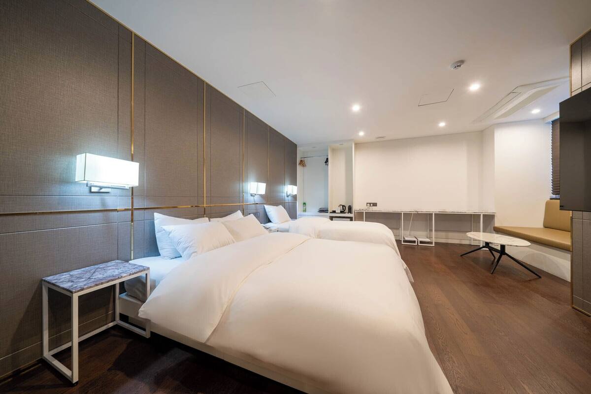 Ein modernes Hotelzimmer mit einem großen Bett, Nachttischen, einem kleinen Schreibtisch und einer Sitzecke mit Tisch und Bank.