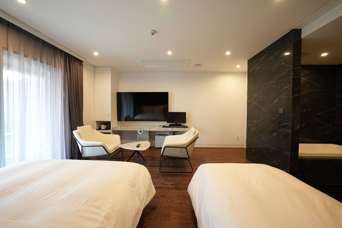 Ein modernes Hotelzimmer mit zwei Betten, einem Schreibtisch mit Flachbildfernseher, einer Sitzecke mit Stühlen und einem großen Spiegel.