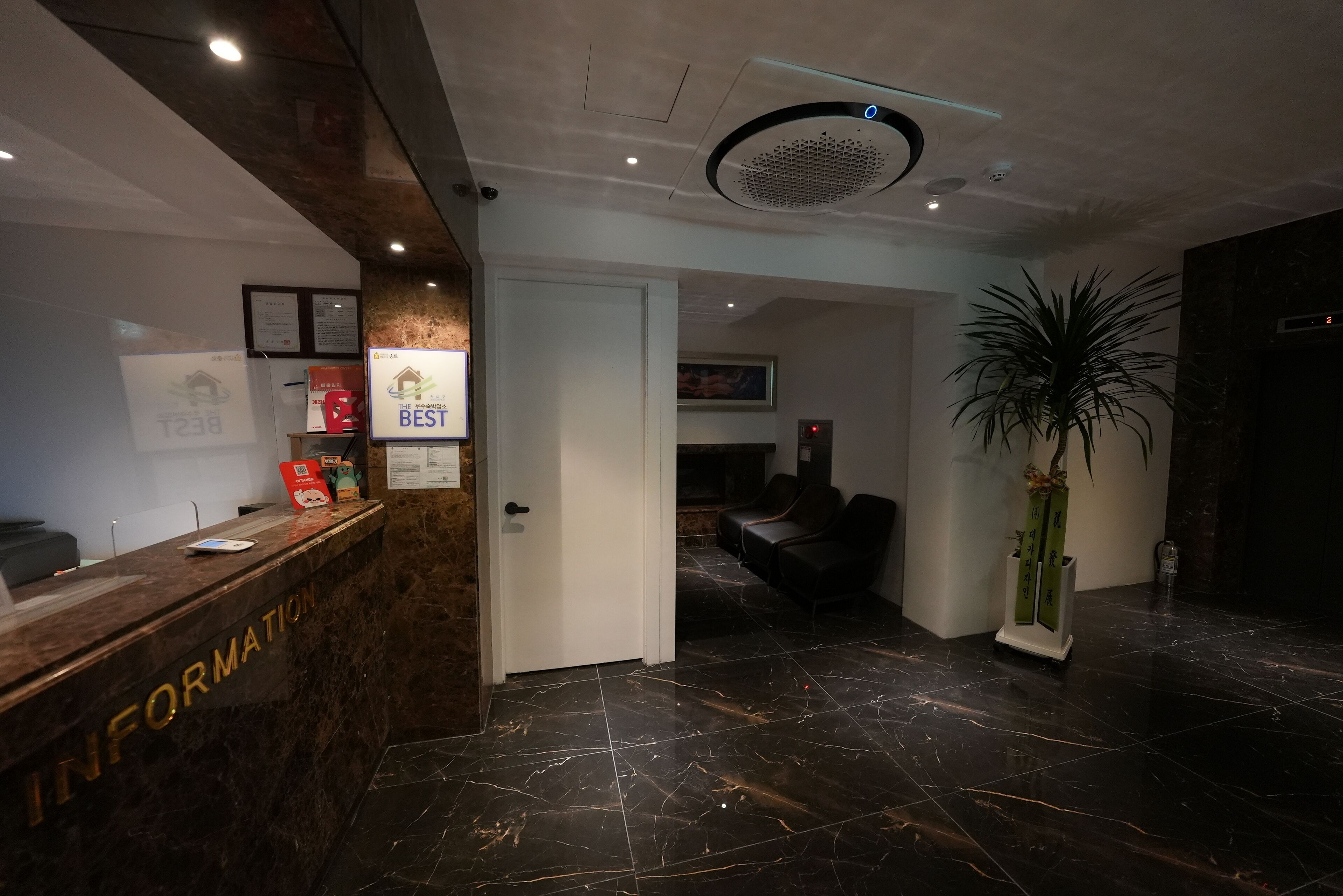 Un espace d’accueil avec un comptoir en marbre portant l’inscription « RENSEIGNEMENTS », une porte donnant sur un espace détente et une plante en pot.