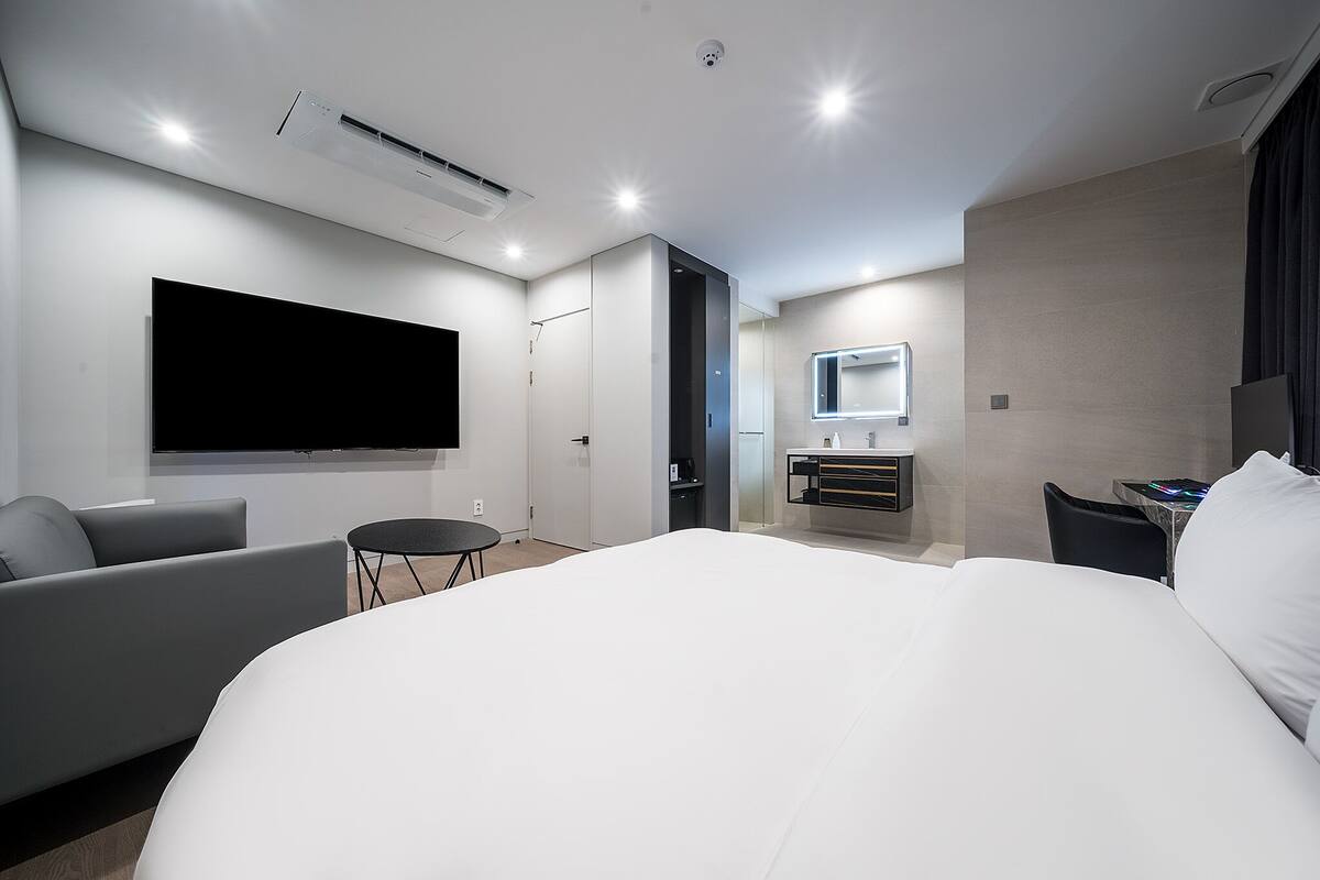 Ein modernes Hotelzimmer mit einem großen Bett, einem flachbildfernseher an der Wand, einem kleinen runden Tisch, einer Couch und einem Schminktisch mit Spiegel.