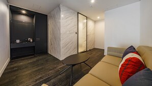 Un interior moderno con piso de madera oscura, un sofá color crema con almohadones coloridos, una pequeña mesa redonda y un mueble oscuro con repisa de vidrio.