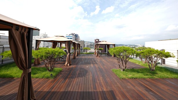 Una terraza de madera con vistas al horizonte de la ciudad, cubierta por toldos y rodeada de árboles pequeños.