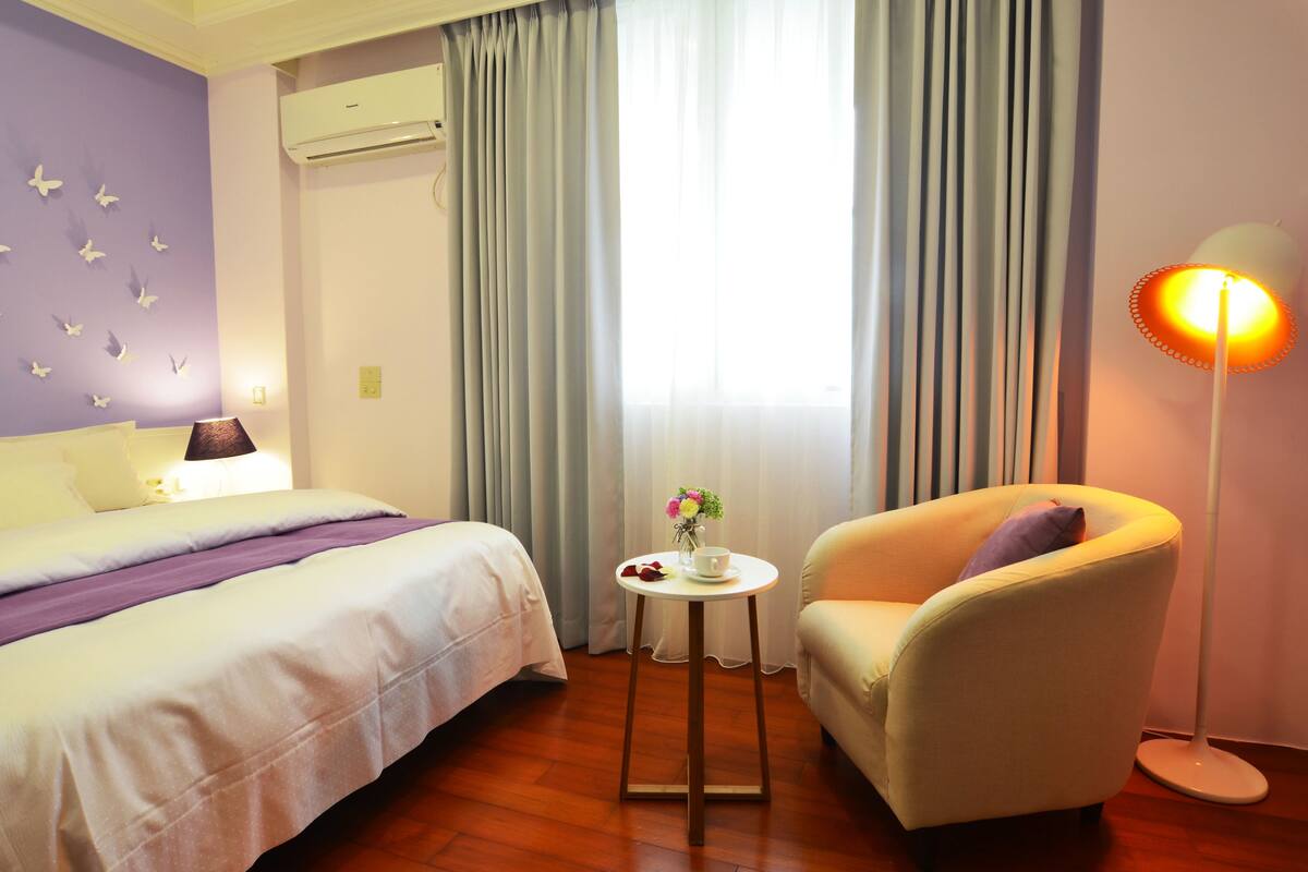 Une chambre d’hôtel avec un lit, une chaise, une petite table sur laquelle se trouve un vase avec une fleur, et un lampadaire.