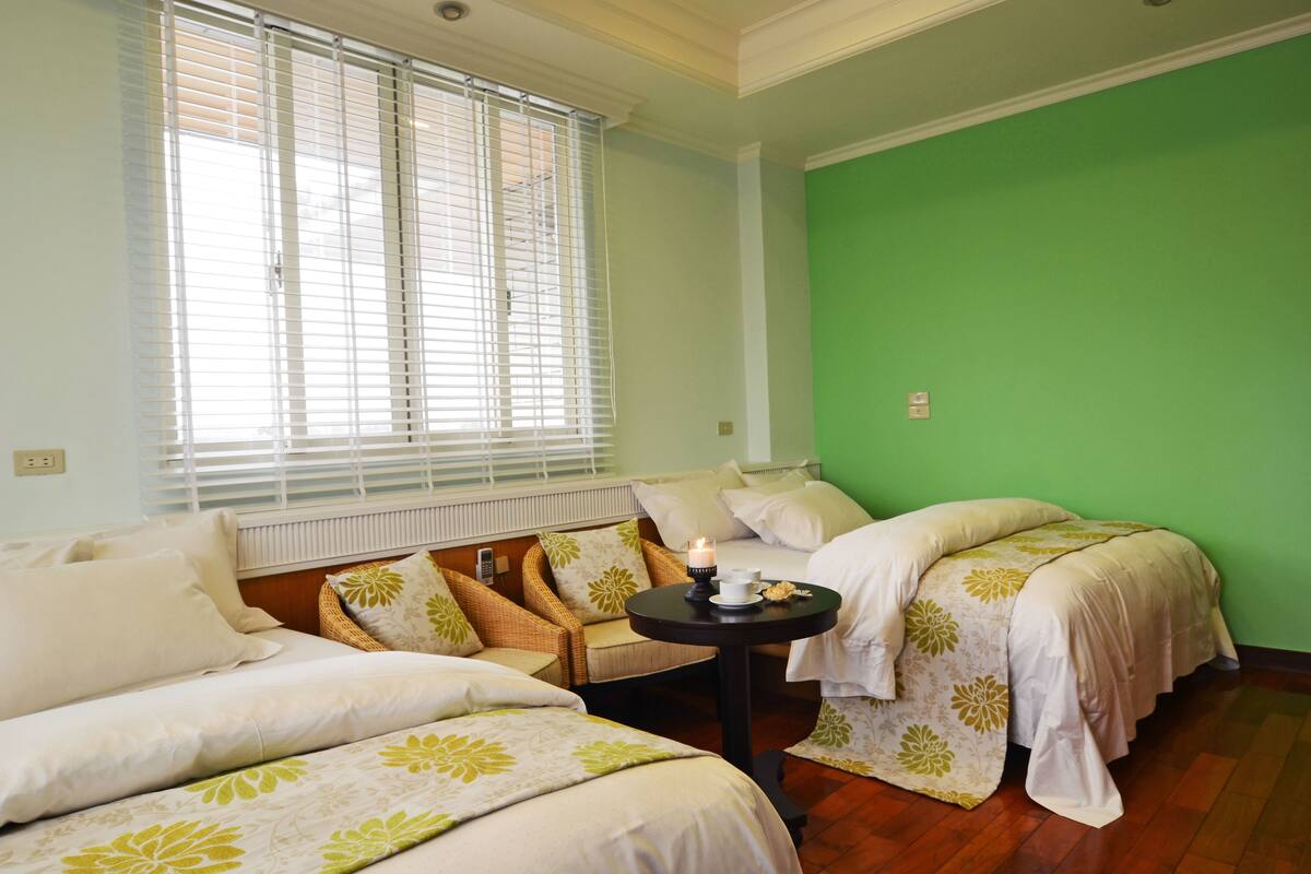 Une chambre avec deux lits, une chaise en osier, une petite table avec une bougie et un mur vert.
