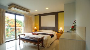 A hotel room with a bed, bedside tables, a bench, a window with curtains, an air conditioning unit, and a view of a balcony with plants. - Diamond City Place (Pattaya)