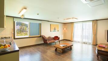 A spacious living room with a wooden floor, a sofa, a coffee table, and a dining table. There is a window with a view of buildings outside.
