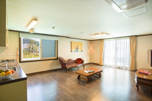 A spacious living room with a wooden floor, a sofa, a coffee table, and a dining table. There is a window with a view of buildings outside.