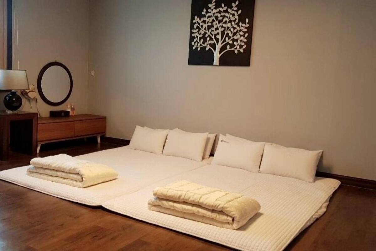 A bedroom with a large bed, two bedside tables, a nightstand, a wall-mounted mirror, and a tree artwork.