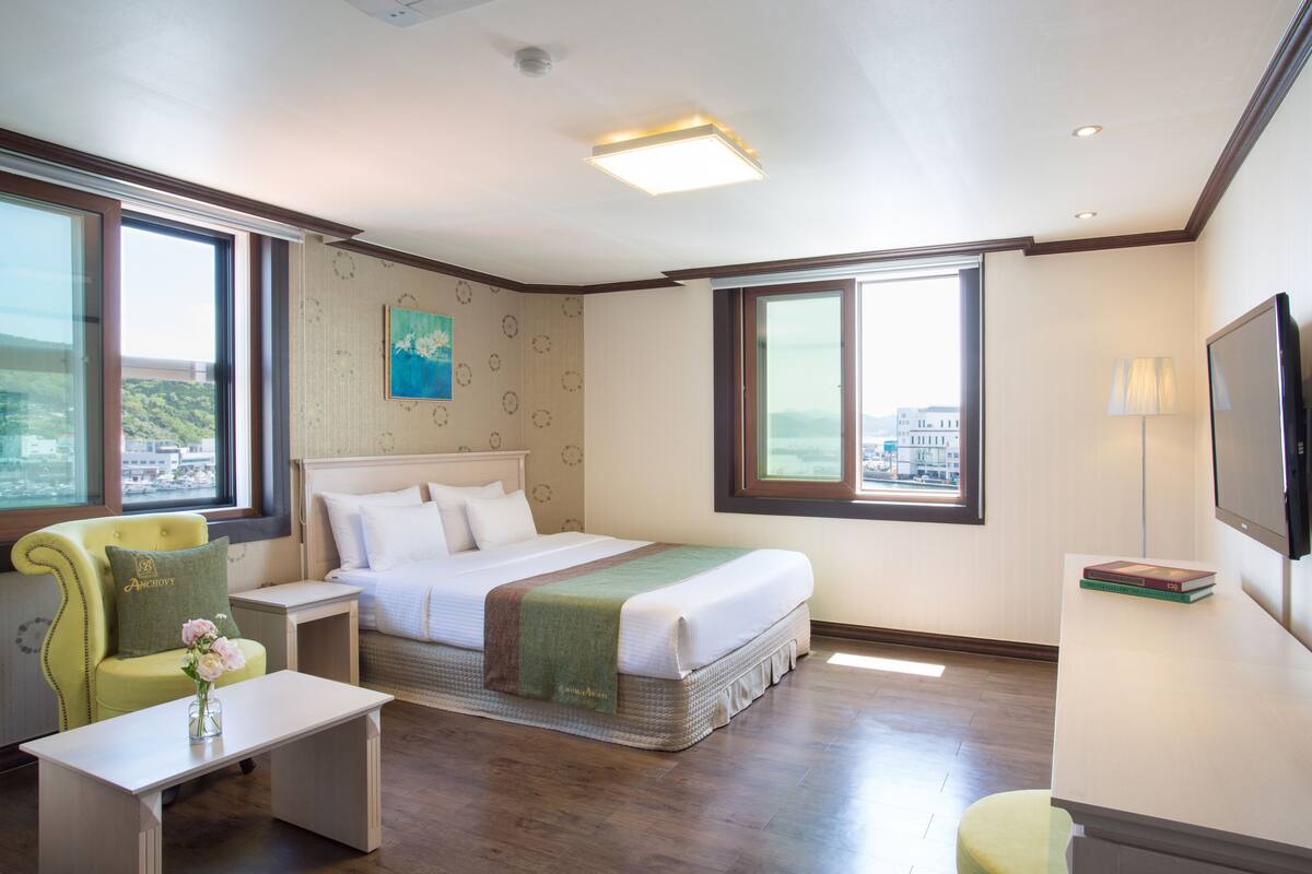 A hotel room with a bed, a chair, a desk, a TV, and large windows offering a city view.