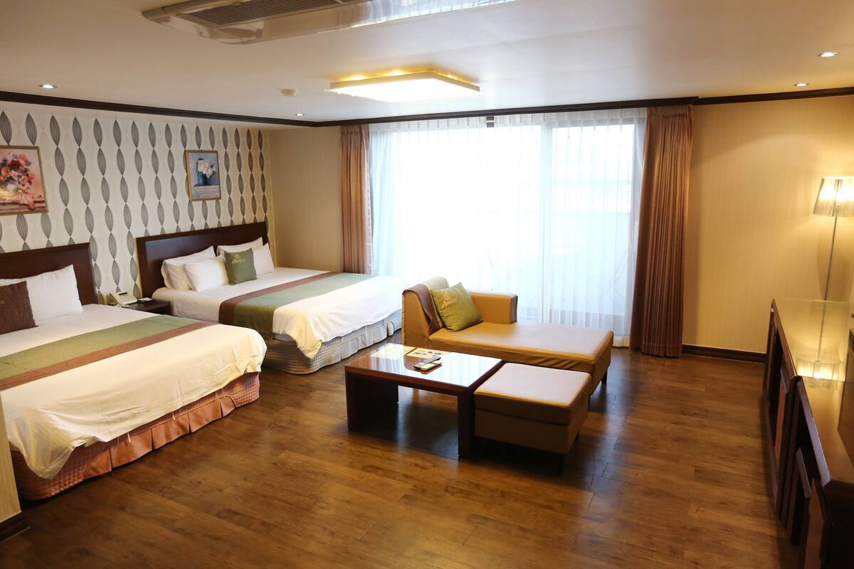 A hotel room with two beds, a sofa, a coffee table, and a large window with curtains.