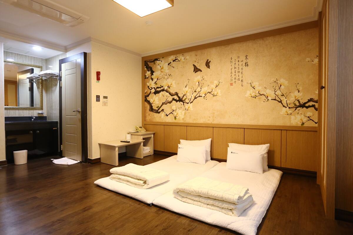 A hotel room with a wooden floor, a large bed with white linens, a wooden headboard, a wall with a floral mural, a small bedside table, and a mirror.