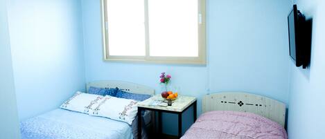 A bedroom with two beds, a window, a bedside table with a vase of flowers, and a wall-mounted TV.